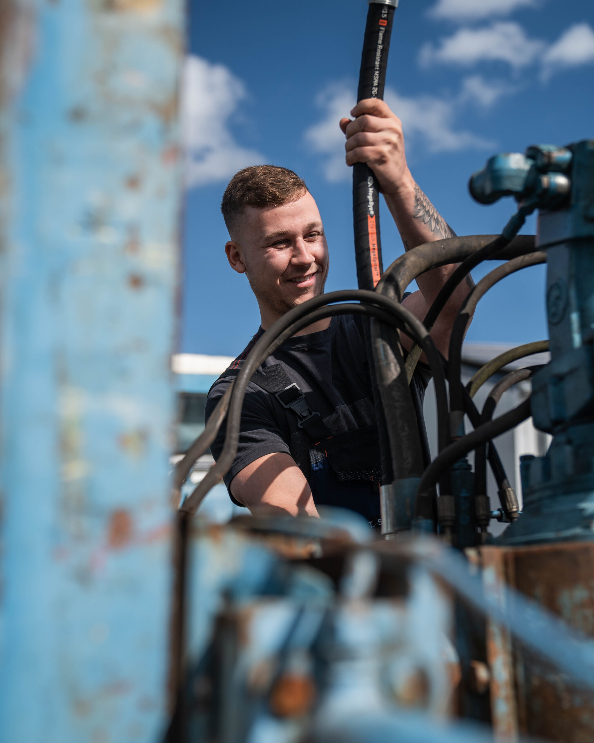 Junger Mann im T-Shirt montiert einen Schlauch in eine Vorrichtung eines Gerätes, das für den Brunnenbau eingesetzt wird. Der Himmel ist blau mit ein paar kleinen Wolken.