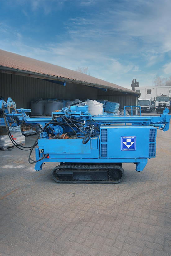 Ein großes, blaues Bohrgerät aus dem Fuhrpark von Ivers Brunnenbau steht frei auf einem gepflasterten Hofplatz. Die Maschine ist auf Ketten montiert. Im Hintergrund ist eine kleine Lagerhalle zu sehen.