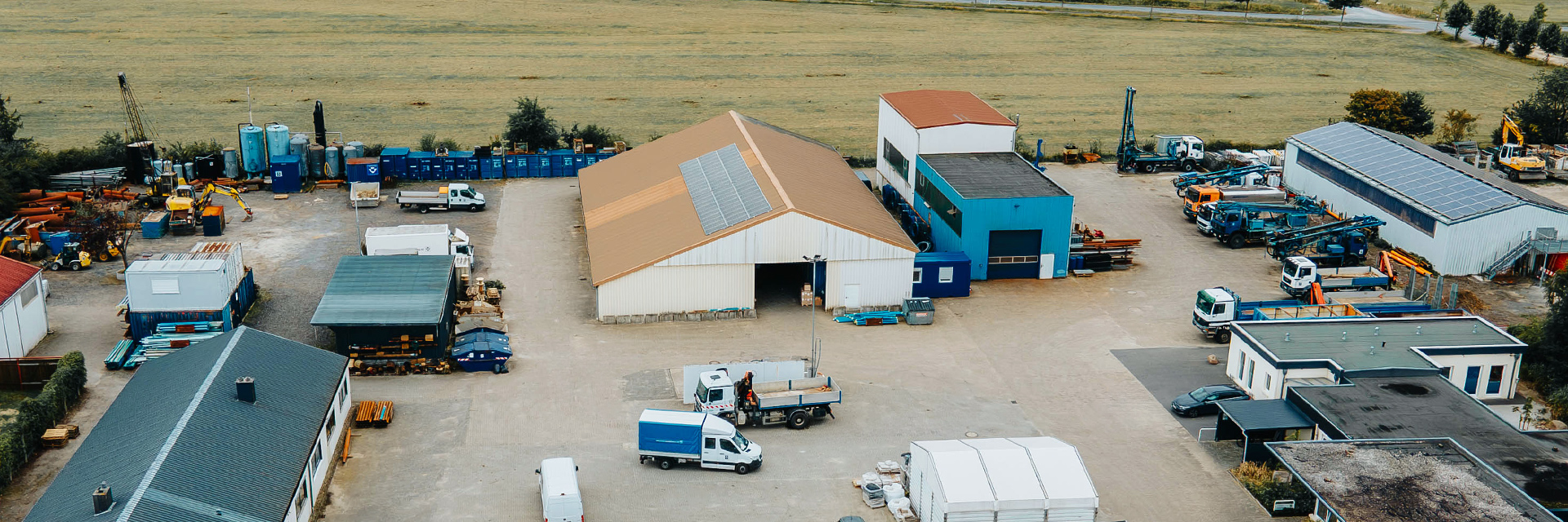 Eine Drohnenaufnahme zeigt aus der Vogelperspektive den Standort von Ivers Brunnenbau. Mittig ist eine weiße Scheine mit roten Dach und Photovoltaik. Rechts stehen viele Fahrzeuge in weißen und blauen Farben. Links wird eine weitere Scheine gezeigt und im Hintergrund ist eine Wiese.