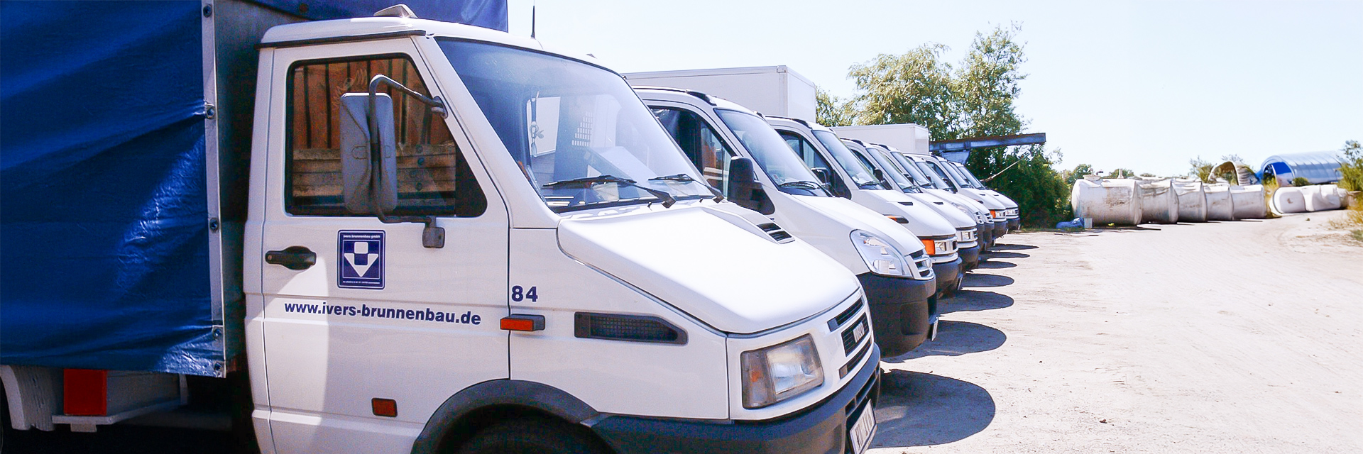 Dieses Bild stellt einen Fuhrpark dar. Mehrere weiße Transporter stehen dicht an dicht in einer Reihe auf einem Betriebshof. Vorne links im Bild ist ein kleiner Lkw mit blauer Plane, auf dessen Tür das Firmenlogo und die Internetadresse ‚ivers-brunnenbau.de‘ zu sehen sind. Rechts am Bildrand liegen große weiße Container, im Hintergrund Bäume und Hallen.