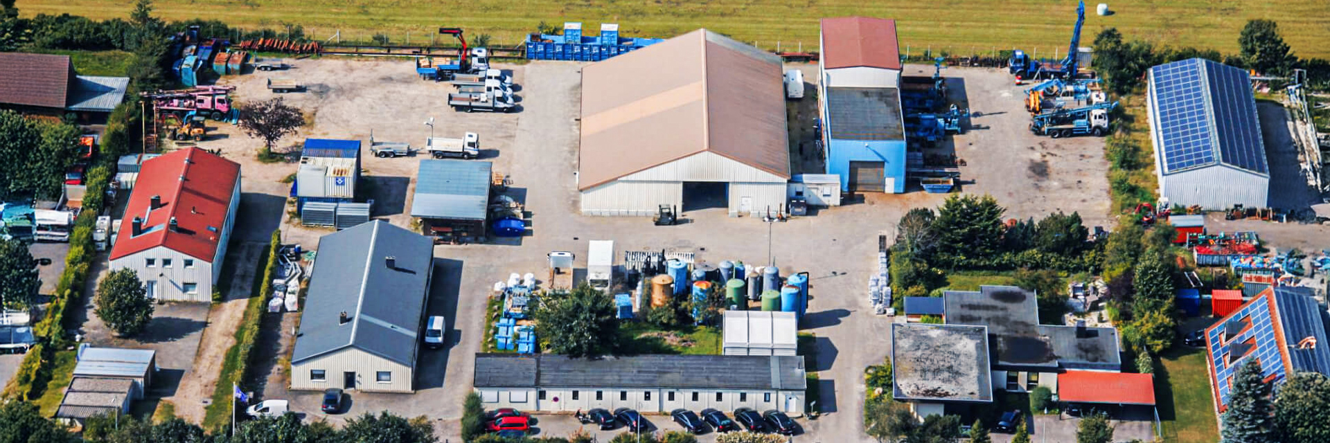 Eine Luftaufnahme zeigt ein großes Firmengelände mit mehreren Hallen, Werkstätten und Lagern. In der Mitte steht eine große Halle mit beigem Dach, daneben Hallen mit blauen und roten Dächern. Auf den Freiflächen sind Fahrzeuge, Container und Tanks abgestellt. Im Hintergrund grenzt das Gelände an Felder.