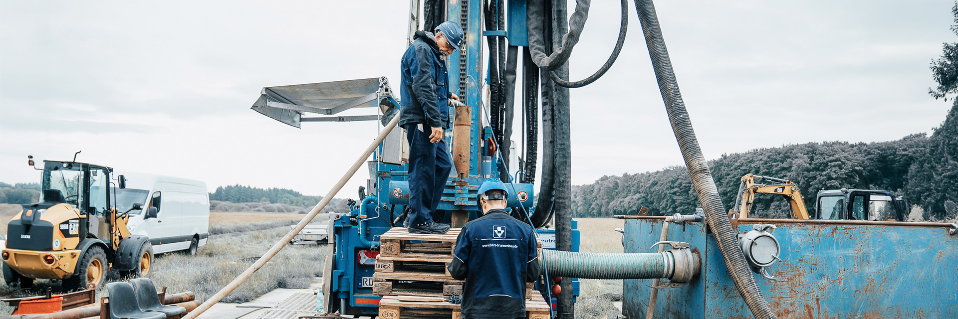 Auf einer offenen Wiese arbeiten zwei Männer in blauer Schutzkleidung an einer großen, senkrecht stehenden Bohrmaschine. Einer steht oben auf Holzpaletten direkt an der Maschine und hantiert mit einem Rohr, der andere steht darunter und schaut nach oben. Rechts führt ein dicker schwarzer Schlauch in einen rostigen Container. Links im Bild stehen ein gelber Radlader und ein weißer Transporter.