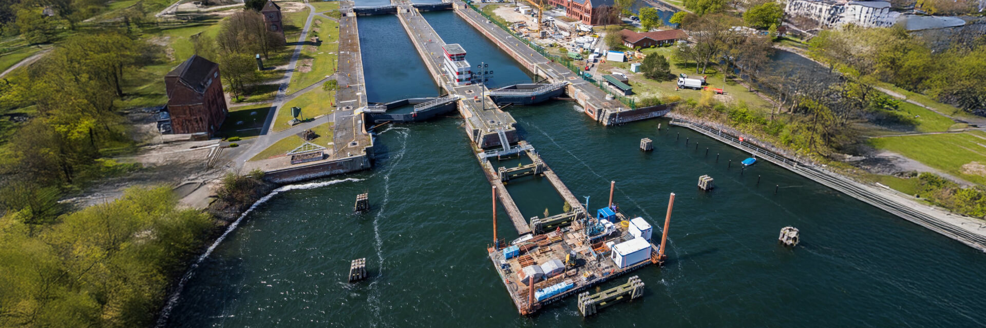 Eine Luftaufnahme zeigt eine große Schleusenanlage an einem Fluss oder Kanal. Es ist das Schlüsselbild für Baugrund. In der Mitte verläuft das Schleusenbecken, das durch massive Mauern eingefasst ist. Im Vordergrund auf dem Wasser befindet sich eine Baustelle mit Kränen, Containern und Maschinen auf einer Plattform. Links und rechts des Kanals sind Grünflächen mit Bäumen, Wegen und Gebäuden zu sehen. Im Hintergrund stehen mehrere rote Backsteingebäude und weitere Baustelleneinrichtungen.