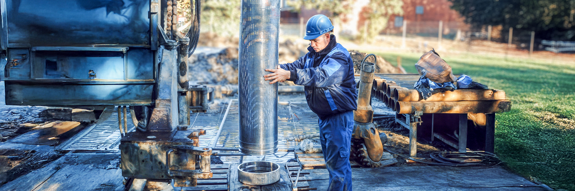 Auf einer Baustelle steht rechts im Bild ein Arbeiter mit blauem Schutzhelm und blauer Arbeitskleidung. Er richtet für den Brunnenbau ein großes, silbrig glänzendes Metallrohr senkrecht über eine runde Öffnung am Boden aus. Links daneben steht ein massives blaues Bohrgerät. Rechts am Bildrand liegen mehrere kurze Rohre auf einer Ablage. Der Boden besteht aus Metallgittern, im Hintergrund sind grüne Wiese und unscharfe Gebäude erkennbar.