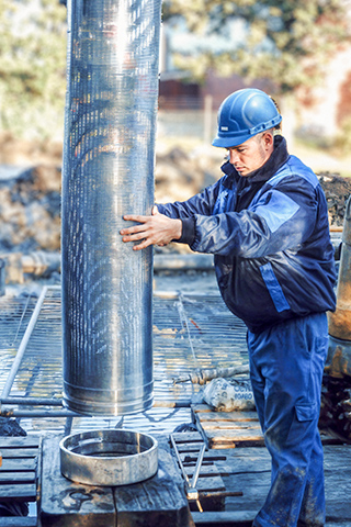 Auf einer Baustelle steht rechts im Bild ein Arbeiter von Ivers Brunnenbau mit blauem Schutzhelm und blauer Arbeitskleidung. Er richtet ein großes, silbrig glänzendes Metallrohr senkrecht über eine runde Öffnung am Boden aus. Links daneben steht ein massives blaues Bohrgerät. Rechts am Bildrand liegen mehrere kurze Rohre auf einer Ablage. Der Boden besteht aus Metallgittern, im Hintergrund sind grüne Wiese und unscharfe Gebäude erkennbar.