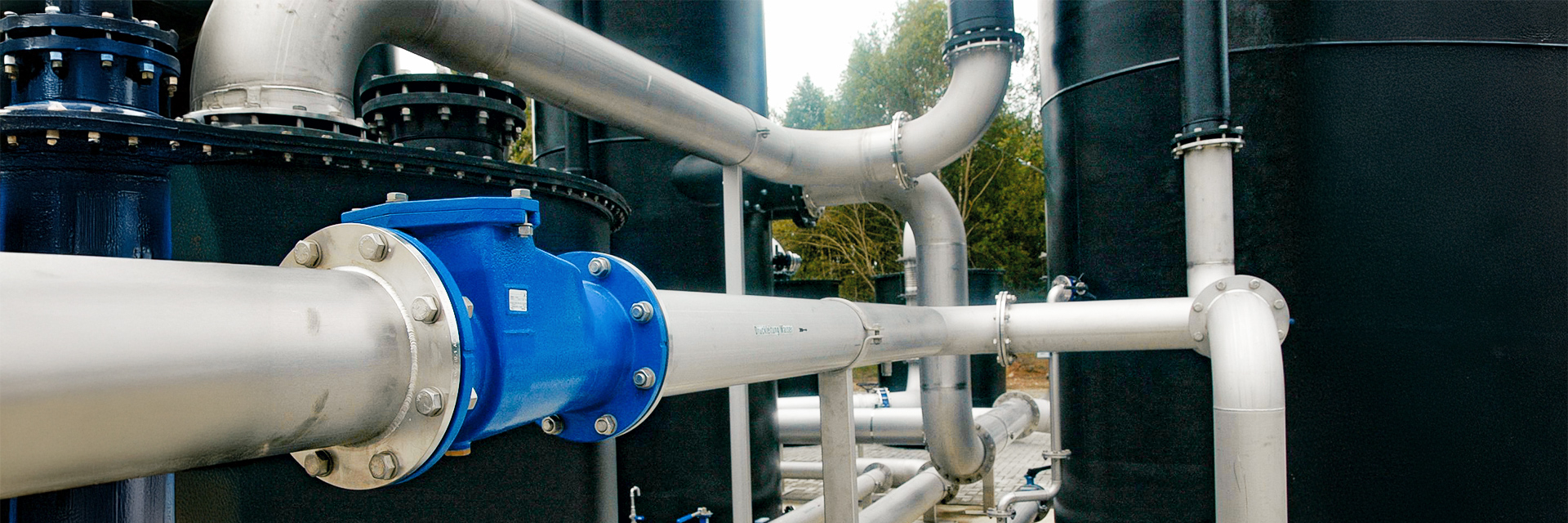 Mehrere breite, silberne Metallrohre verlaufen waagerecht und senkrecht durch die Anlage. Sie sind mit großen Schraubflanschen und Ventilen verbunden, vorne im Bild mit einem blau lackierten Anschlussstück. Im Hintergrund stehen große, schwarze Zylinderanlagen, dahinter sind Bäume zu erkennen.