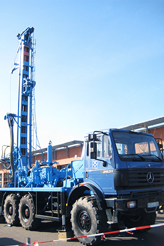 Ein großer blauer Mercedes-Lkw mit Allradreifen aus dem Fuhrpark von Ivers Brunnenbau steht im Freien. Auf der Ladefläche ist eine Bohranlage mit hohem Mast aufgebaut. Das Fahrzeug ist von rot-weißen Absperrbändern umgeben, im Hintergrund Gebäude.