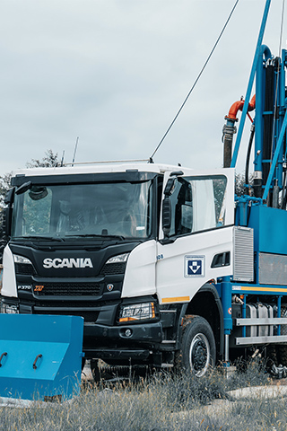 Ein weißer Scania-Lastwagen aus dem Fuhrpark von Ivers Brunnenbau steht schräg nach rechts. Auf der Ladefläche ist ein großes blaues Bohrgerät montiert. Am Heck verlaufen dicke Schläuche nach oben. Links im Bild eine blaue Metallabdeckung, im Hintergrund Bäume und grauer Himmel.