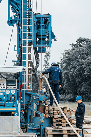 Seitliche Ansicht eines großen blauen Bohrgeräts. Links sind Steuerpulte mit vielen Anzeigen und Knöpfen angebracht. Rechts steht ein Arbeiter auf gestapelten Paletten und hält ein dickes Rohr, während ein Kollege mit Helm am Boden danebensteht. Im Hintergrund Bäume und grauer Himmel.