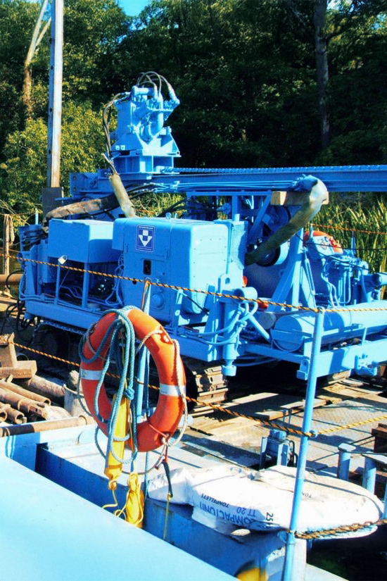 Ein großes, blaues Bohrgerät aus dem Fuhrpark von Ivers Brunnenbau steht auf einem Floß. Die Maschine ist auf Ketten montiert. Im Vordergrund ist ein orangener Rettungsring. Im Hintergrund sind ein Mast und viele grüne Bäume.