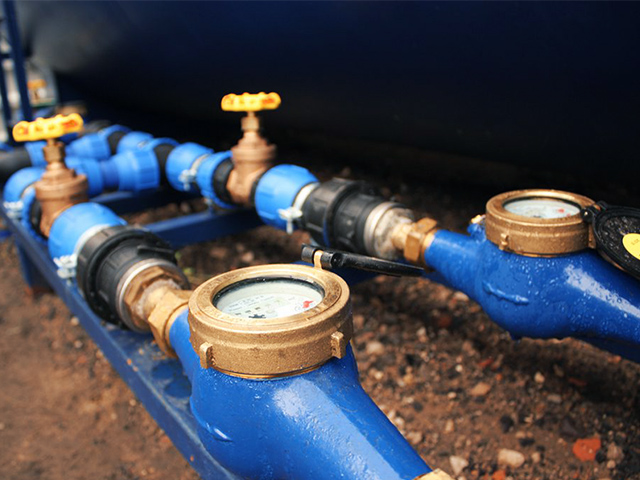 Im Vordergrund sind blaue Metallrohre mit mehreren Wasseruhren und Ventilen sichtbar. Die Uhren sind rund und mit Glas abgedeckt, die Ventile sind mit gelben Handrädern versehen. Die Rohre verlaufen nebeneinander und sind mit schwarzen Kunststoffverbindungen gekoppelt. Darunter liegt grober Kiesboden.