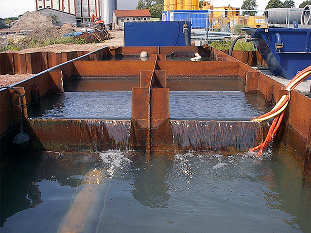 Mehrere rechteckige Becken aus rostbraunem Metall sind mit Wasser gefüllt. In der Mitte läuft das Wasser in einem gleichmäßigen Schwall über eine Kante in das vordere Becken. Rechts führen dicke orangefarbene Schläuche hinein. Im Hintergrund stehen Baucontainer, gelbe Silos und Maschinen auf einem Firmengelände.
