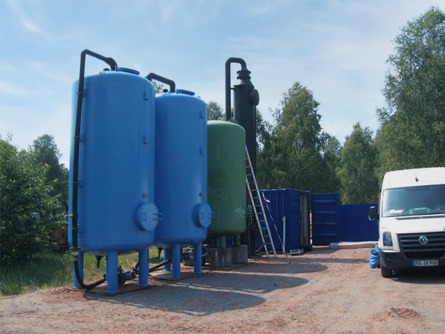 Im Freien stehen drei große, aufrecht stehende Zylindertanks, zwei blau und einer grün. Sie sind über Rohre miteinander verbunden. Rechts daneben ist eine hohe, schwarze Filteranlage mit angeschlossener Leiter. Im Hintergrund stehen blaue Container und Bäume, rechts im Bild ein weißer Transporter.