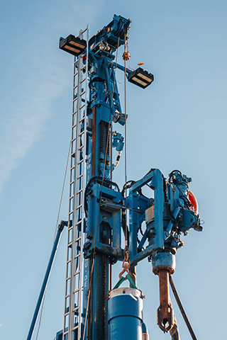 Blick von unten auf eine sehr hohe blaue Maschine. An einem Haken hängt ein großes zylindrisches Metallteil, das mit Gurten befestigt ist. Leitern, Schläuche und Metallstreben verlaufen entlang der Konstruktion.