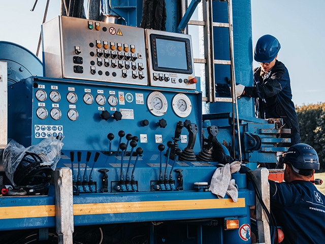 Großes blaues Bedienfeld mit vielen Knöpfen, Hebeln und runden Anzeigen. Zwei Personen mit Schutzhelmen arbeiten daran: eine steht seitlich an der Maschine, eine weitere ist von hinten zu sehen und greift an das Bedienfeld.