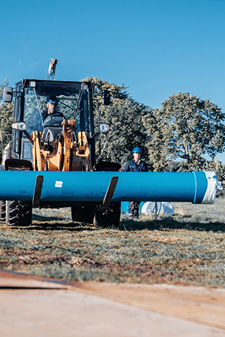 Ein großes Baufahrzeug transportiert ein langes blaues Rohr knapp über dem Boden. Eine Person sitzt im Fahrzeug, eine weitere steht daneben. Im Hintergrund sind Bäume und eine Wiese zu sehen
