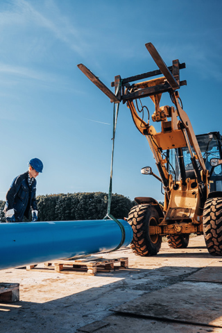 Eine Person mit Schutzhelm steht neben einem langen blauen Rohr, das mit Gurten angehoben wird. Daneben befindet sich ein großes gelbes Baufahrzeug mit breiten Reifen. Die Szene spielt auf einer Baustelle im Freien.