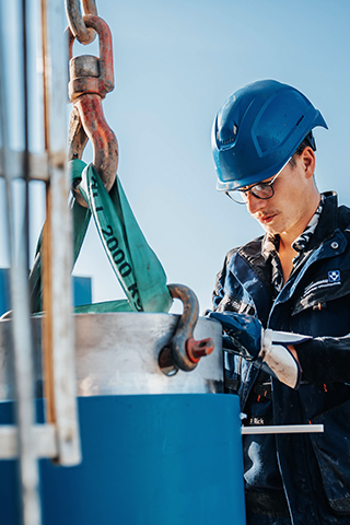 Eine Person mit Schutzhelm und Handschuhen arbeitet konzentriert an einem großen blauen Metallteil, das an einem Haken hängt. Sie hält ein Werkzeug in den Händen und blickt nach unten auf ihre Arbeit. Die Szene spielt im Freien.