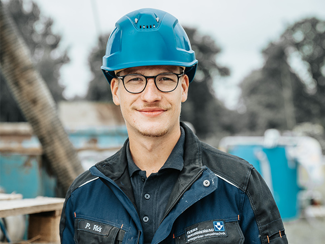 Ein junger Mann mit Brille und blauem Schutzhelm schaut lächelnd in die Kamera. Er trägt eine dunkelblaue Arbeitsjacke mit Namensschild und Firmenlogo. Im unscharfen Hintergrund sind Baumaschinen und Rohre zu erkennen.