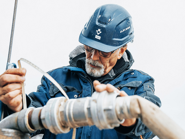 Ein älterer Mann mit grauem Bart und Schutzhelm arbeitet konzentriert an einer großen Rohrverbindung. Er trägt eine dunkelblaue Jacke und hält mit beiden Händen Schläuche und ein Messband. Der Hintergrund ist hell und unscharf.